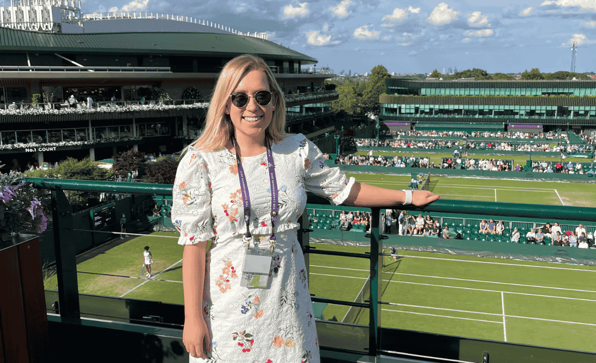 A person in a floral dress stands on a balcony overlooking a tennis court at a well-attended tournament. The scene includes a green stadium and clear blue skies.