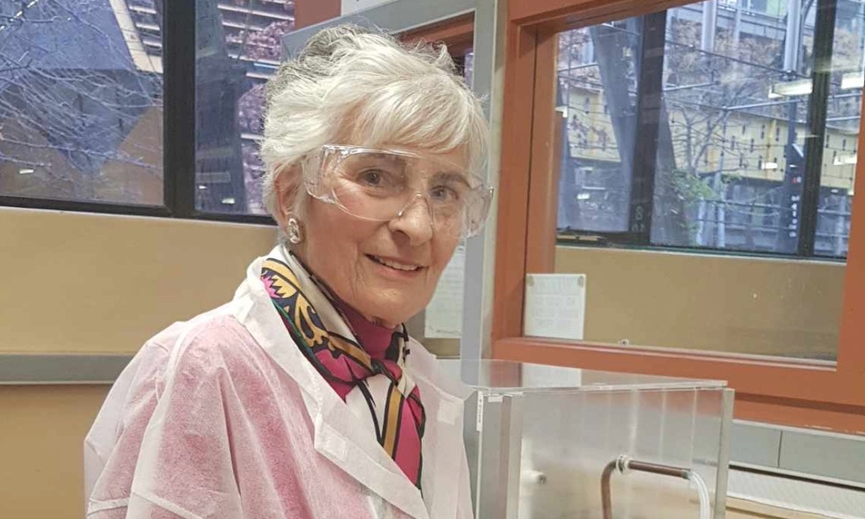 A woman with short white hair stands smiling in a lab, wearing safety glasses, a lab coat and a colourful scarf. She is Emeritus Professor Felicity Roddick, visiting the RMIT wastewater treatment lab working on fatberg prevention.