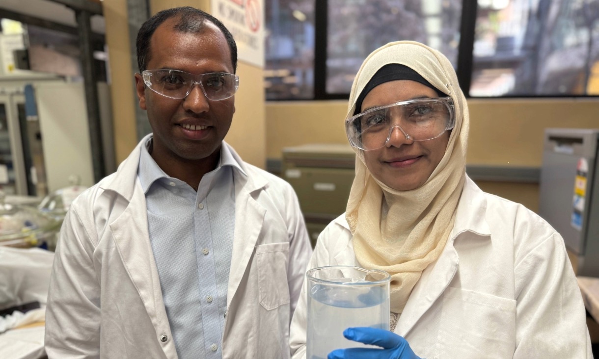 Two researchers, a man and a woman, smile at the camera in a lab. Both wear lab coats, safety glasses and gloves. The woman holds a large beaker filled with clear water. They are Dr Biplob Pramanik and Dr Nilufa Sultana of RMIT University.