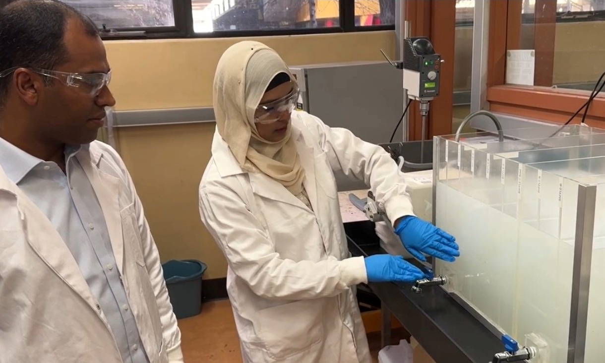 Two engineers in lab coats and safety glasses stand beside a transparent grease interceptor system. The woman, wearing a hijab and blue gloves, explains the setup. The man watches attentively. They are conducting research on wastewater treatment technology at RMIT University.