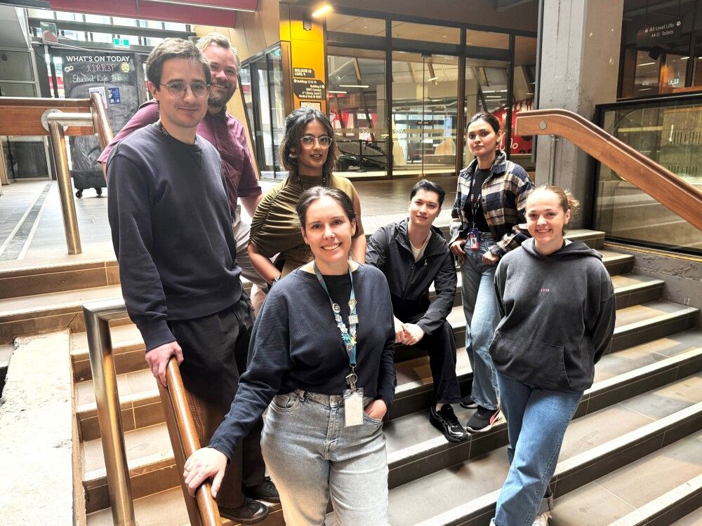 Seven researchers standing and sitting on stairs in an indoor campus setting.