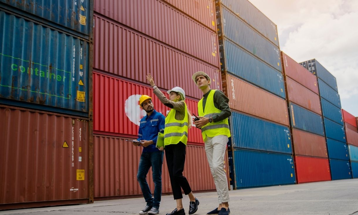 Three people walking along port surrounded by shipping containers