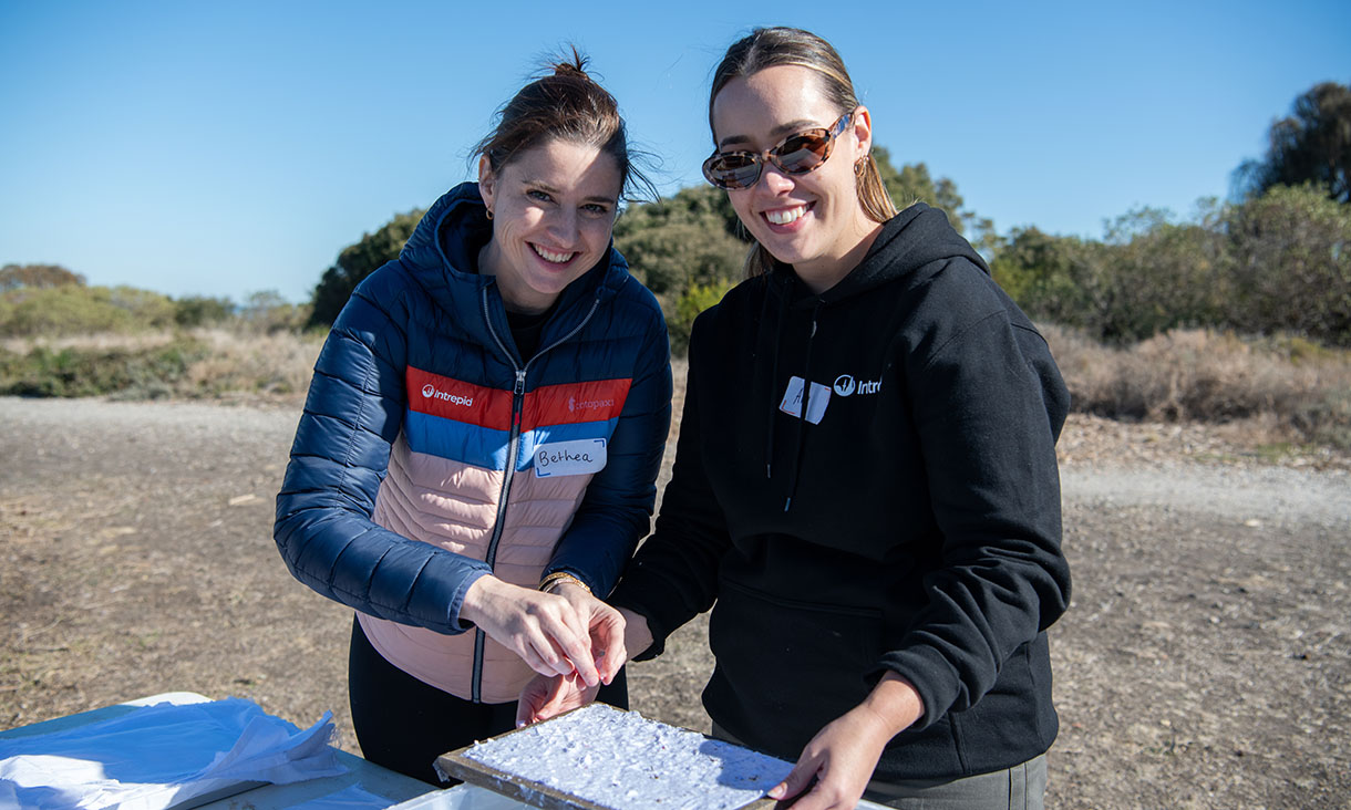 Two people are working together, focused on a tray, set against a natural outdoor background.