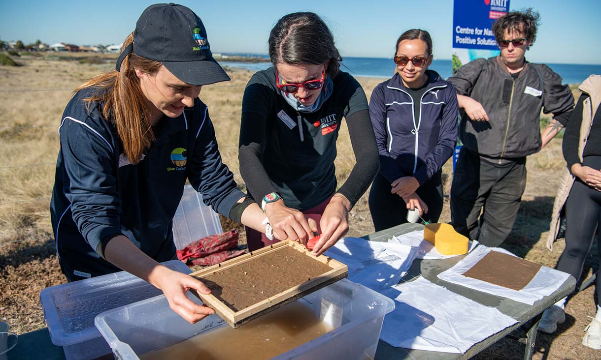 Several people engaged in an outdoor activity, using a wooden frame over a container filled with water and pulp.