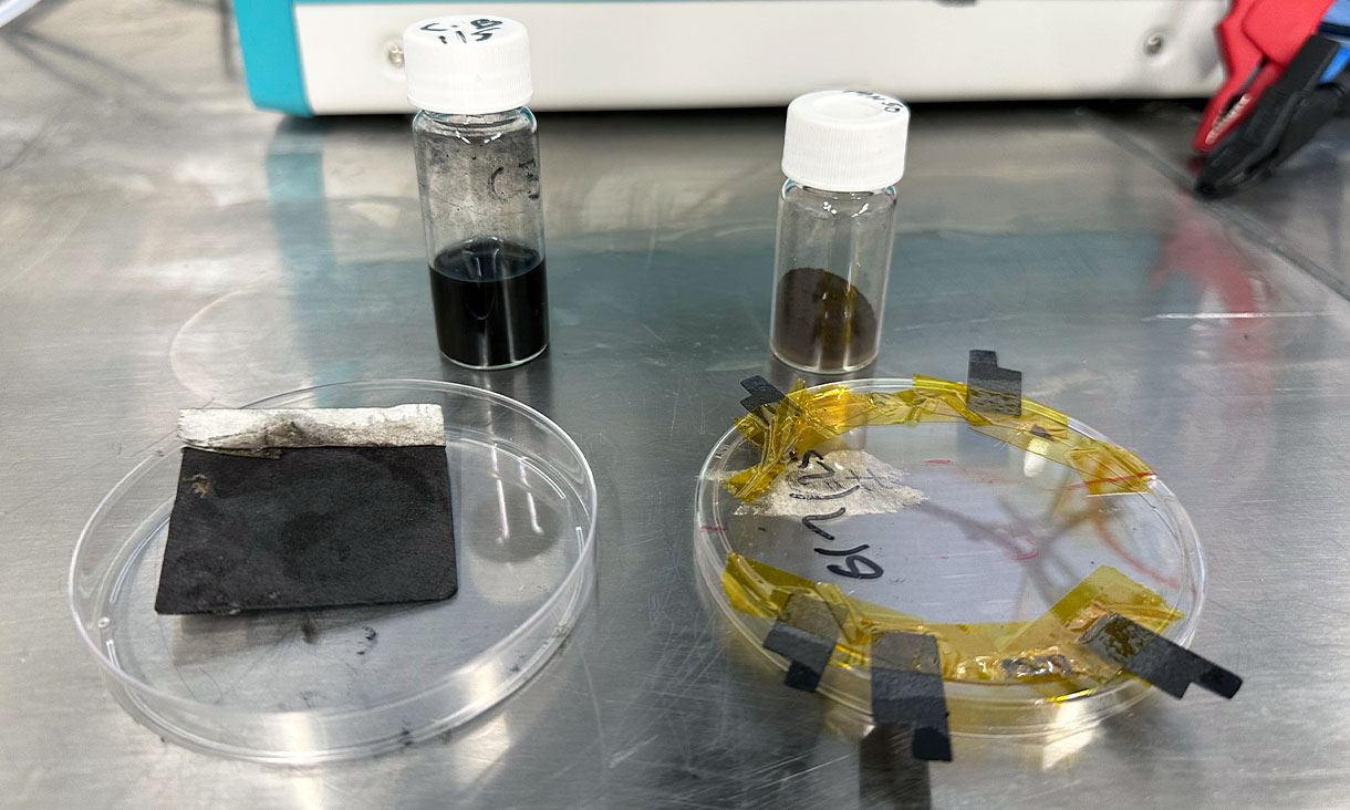 Two petri dishes on a metal benchtop with black strips attached to them, along with two vials of dark coloured substances.