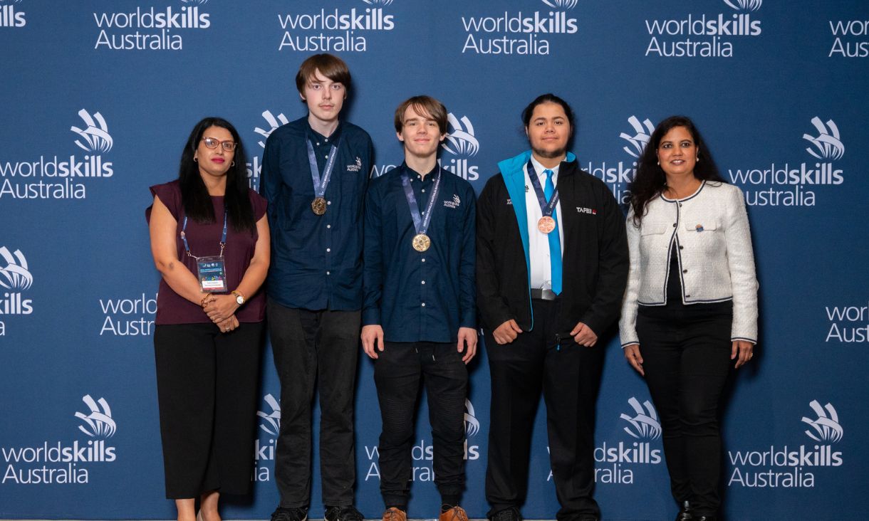 Three competition winners with gold, silver and bronze medals, standing next to two smartly dressed women.
