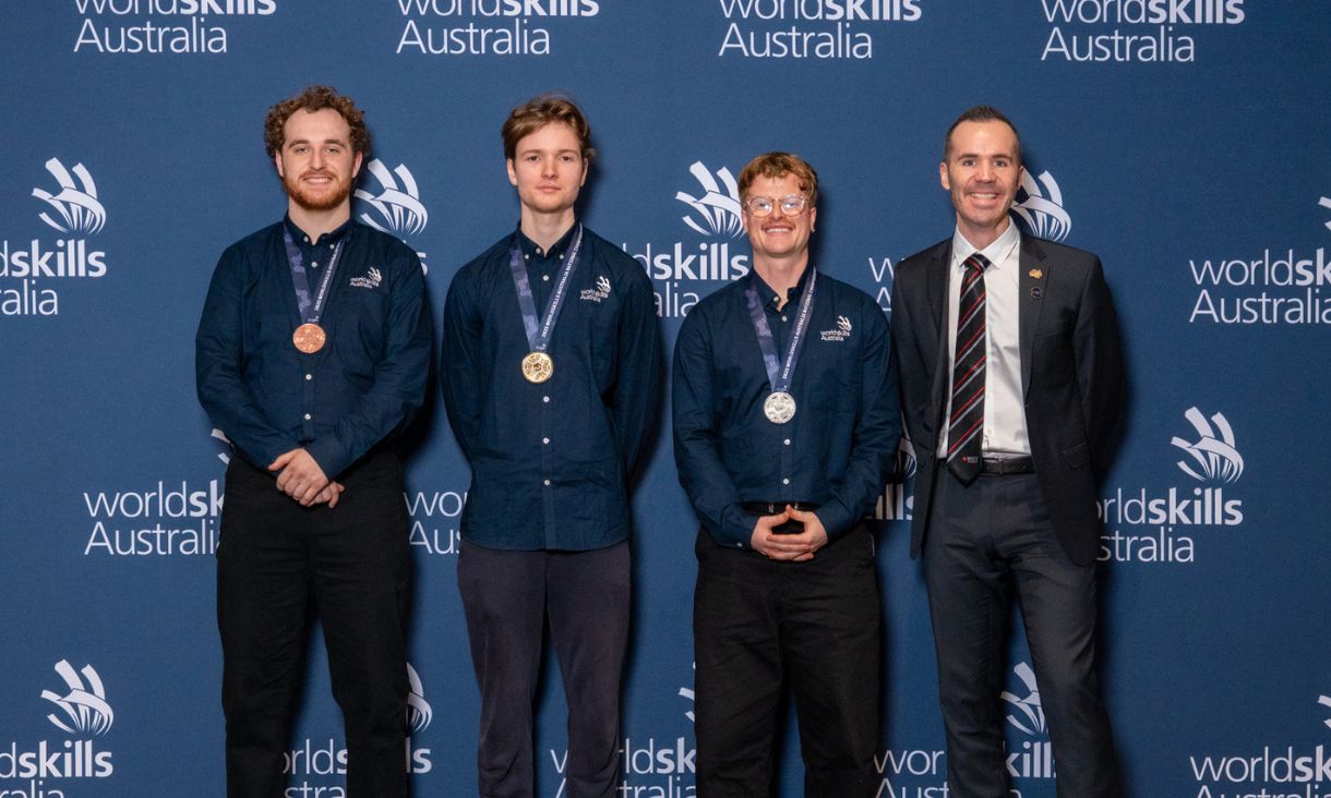 Three competition winners with gold, silver and bronze medals, standing next to man in suit.