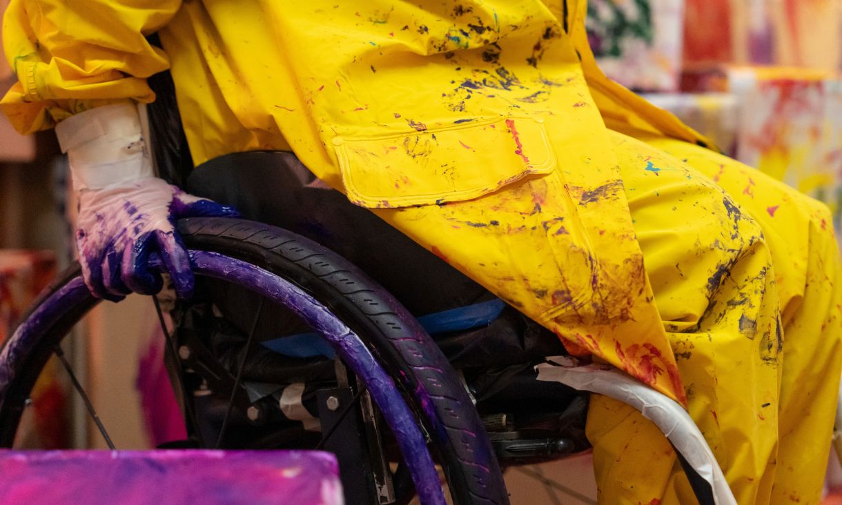 Close up of person in yellow smock sitting in wheelchair with purple paint on its wheels