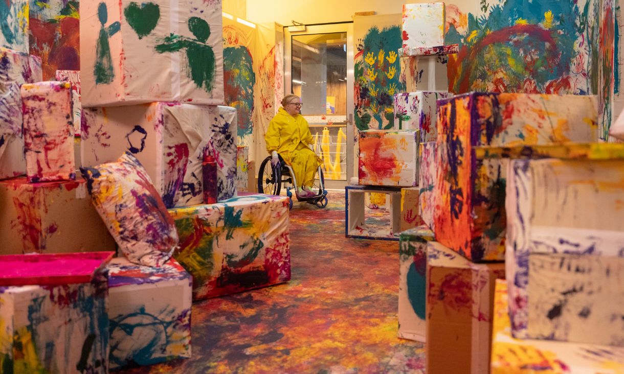 Person in wheelchair in room surrounded by objects that have been splattered with colourful paint