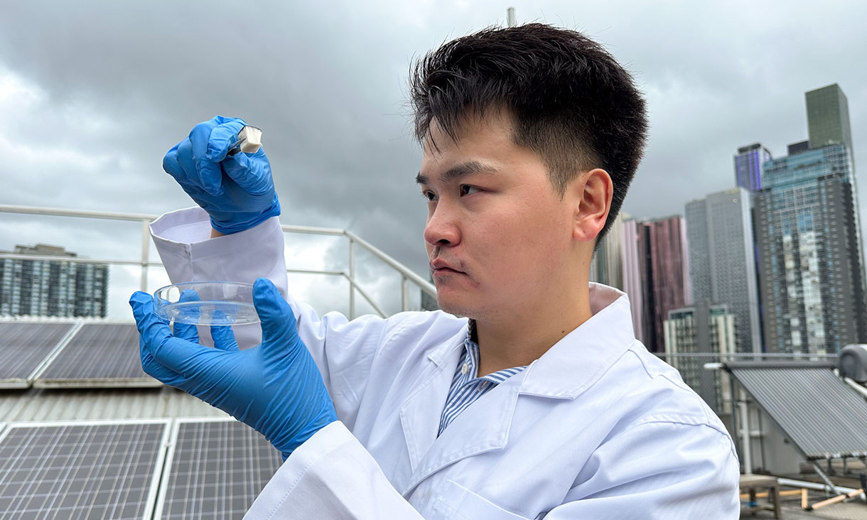 A man wearing blue gloves and a white lab coat looks at a small object in his right hand.