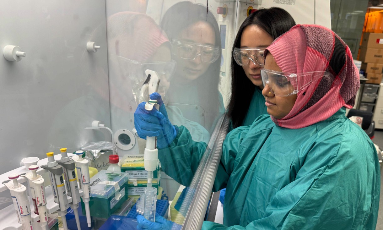 Dr Baoyue Zhang and Dr Sanjida Afrin, wearing lab gowns, gloves and safety glasses, work together at a fume hood while handling laboratory pipettes.