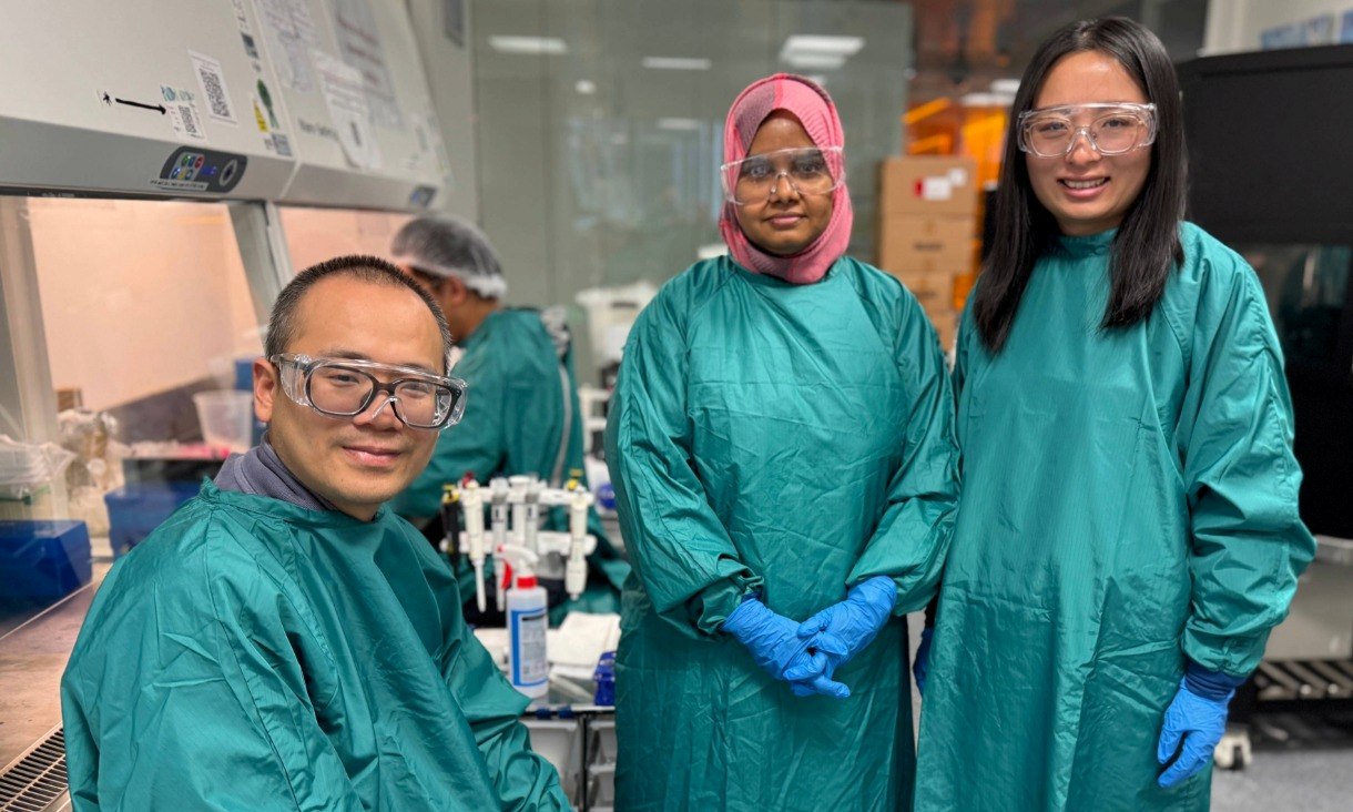 Professor Jianzhen Ou, Sanjida Afrin and Dr Baoyue Zhang wear teal protective gowns, gloves and safety glasses while standing in a laboratory workspace.