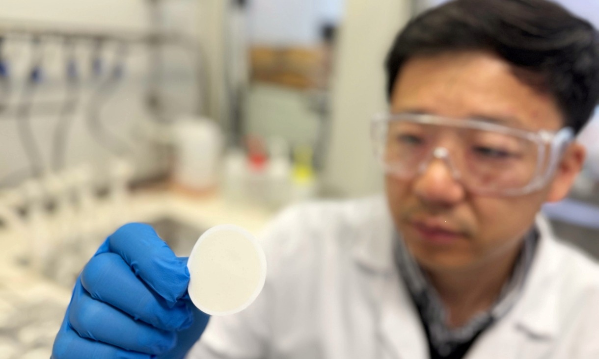 A researcher wearing safety glasses and blue gloves holds a small circular white filter in a laboratory. The background is softly blurred with lab equipment visible.