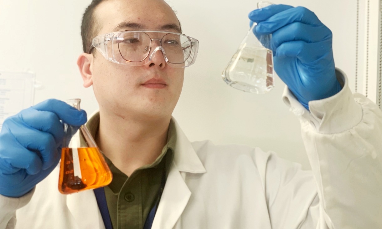 A researcher wearing safety glasses, a white lab coat and blue gloves holds up two glass flasks – one containing a clear liquid and the other an orange solution – examining them closely in a laboratory.