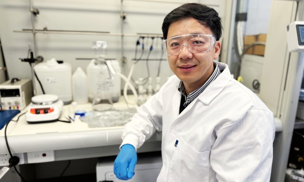 A researcher wearing a white lab coat, blue gloves and safety glasses sits at a laboratory bench with scientific equipment and bottles in the background, smiling at the camera.