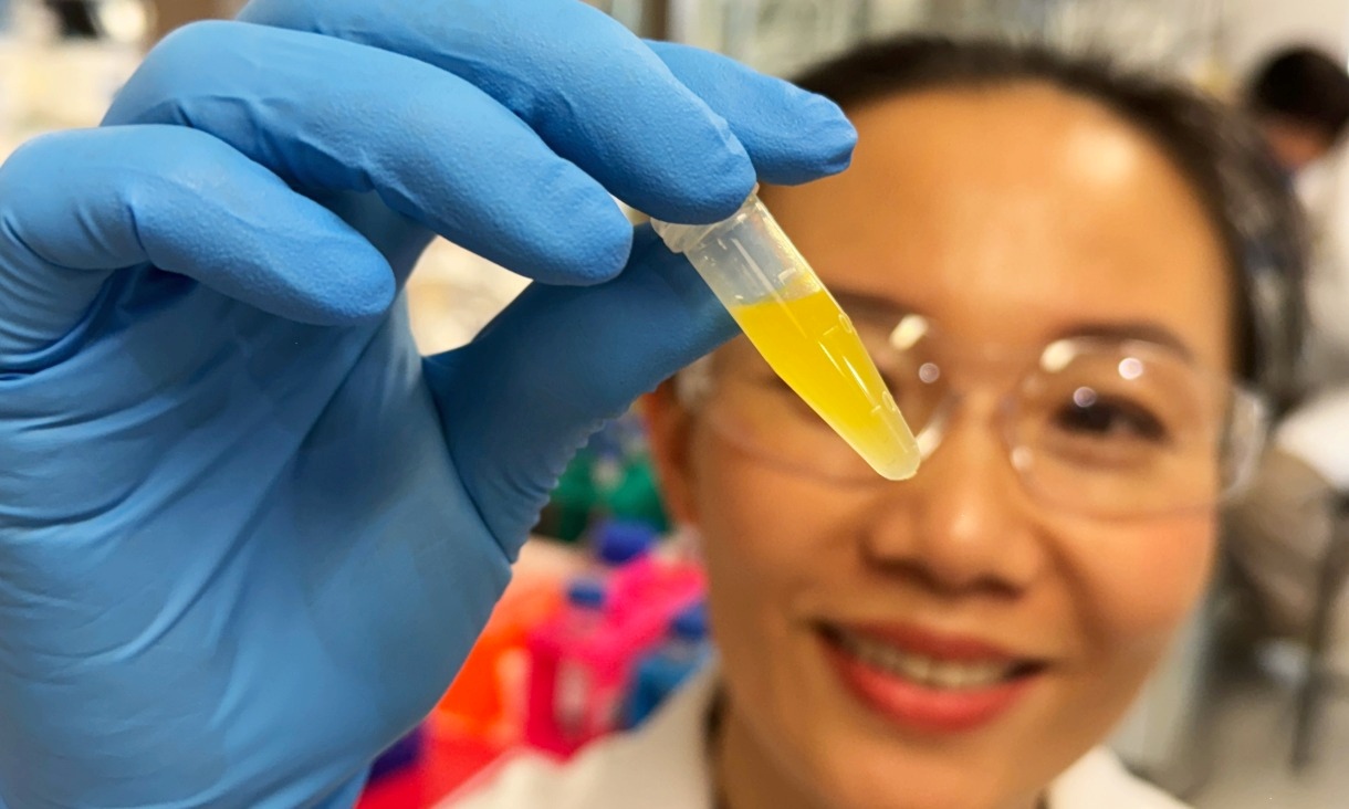 A woman in a lab coat and safety glasses holds up a small vial containing an orange liquid in a laboratory.