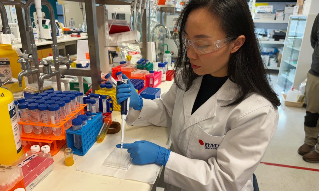 A woman in a white RMIT lab coat and safety glasses sits at a laboratory bench pipetting an orange liquid into a clear plate surrounded by racks of test tubes.