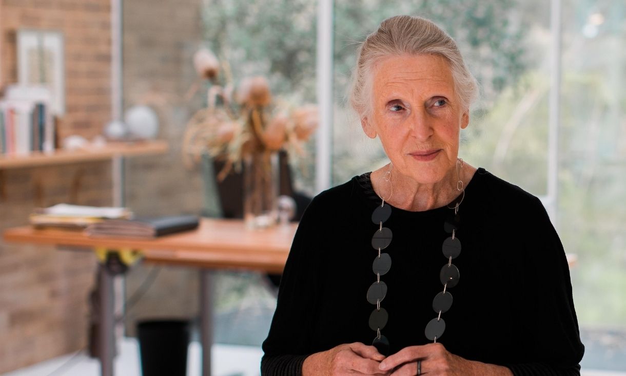Mary stands in front of a large room with a spacious window, she is wearing black with a long necklace.