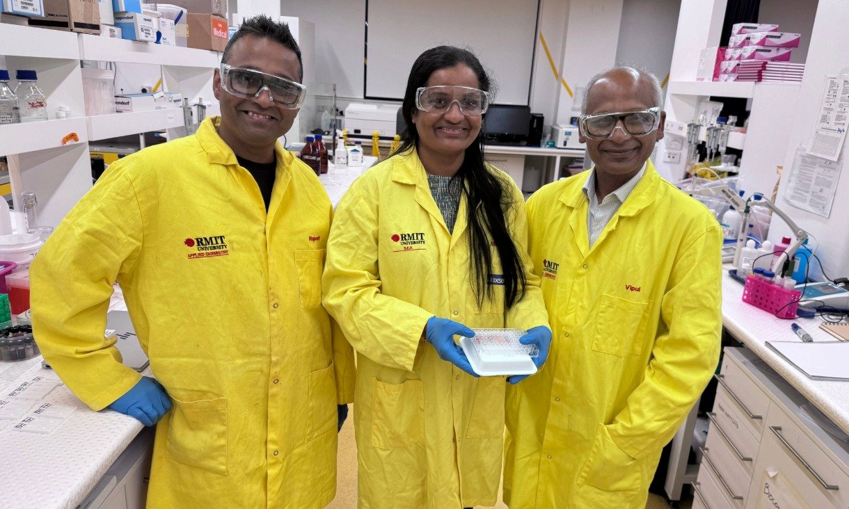 Three RMIT researchers wearing yellow lab coats and safety glasses stand side by side in a laboratory. The researcher in the centre holds a multi well plate used for colour-based golden staph testing, with laboratory benches and equipment behind them.