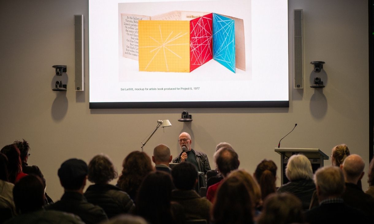 John Kaldor sits in front of a projector screen in font of an audience - screen shows folded paper in blue, yellow and red.