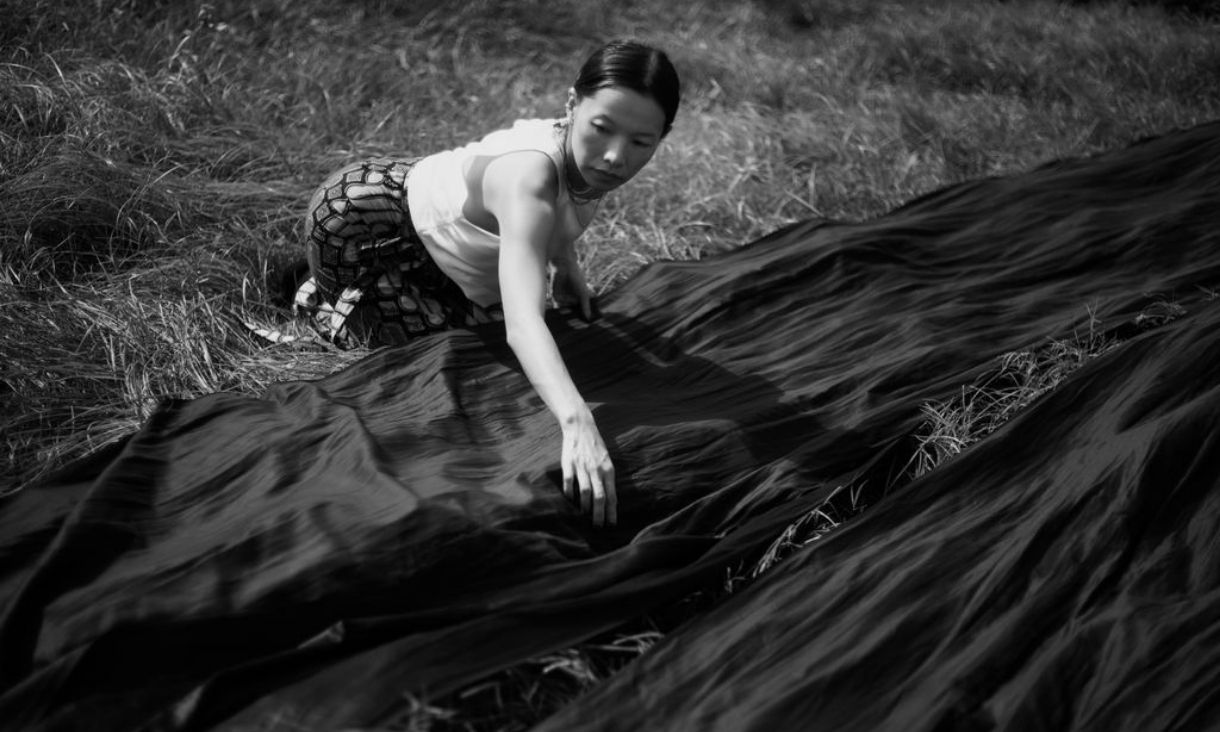 A black and white image of a woman laying sark silk over grass, as if to dry it. 