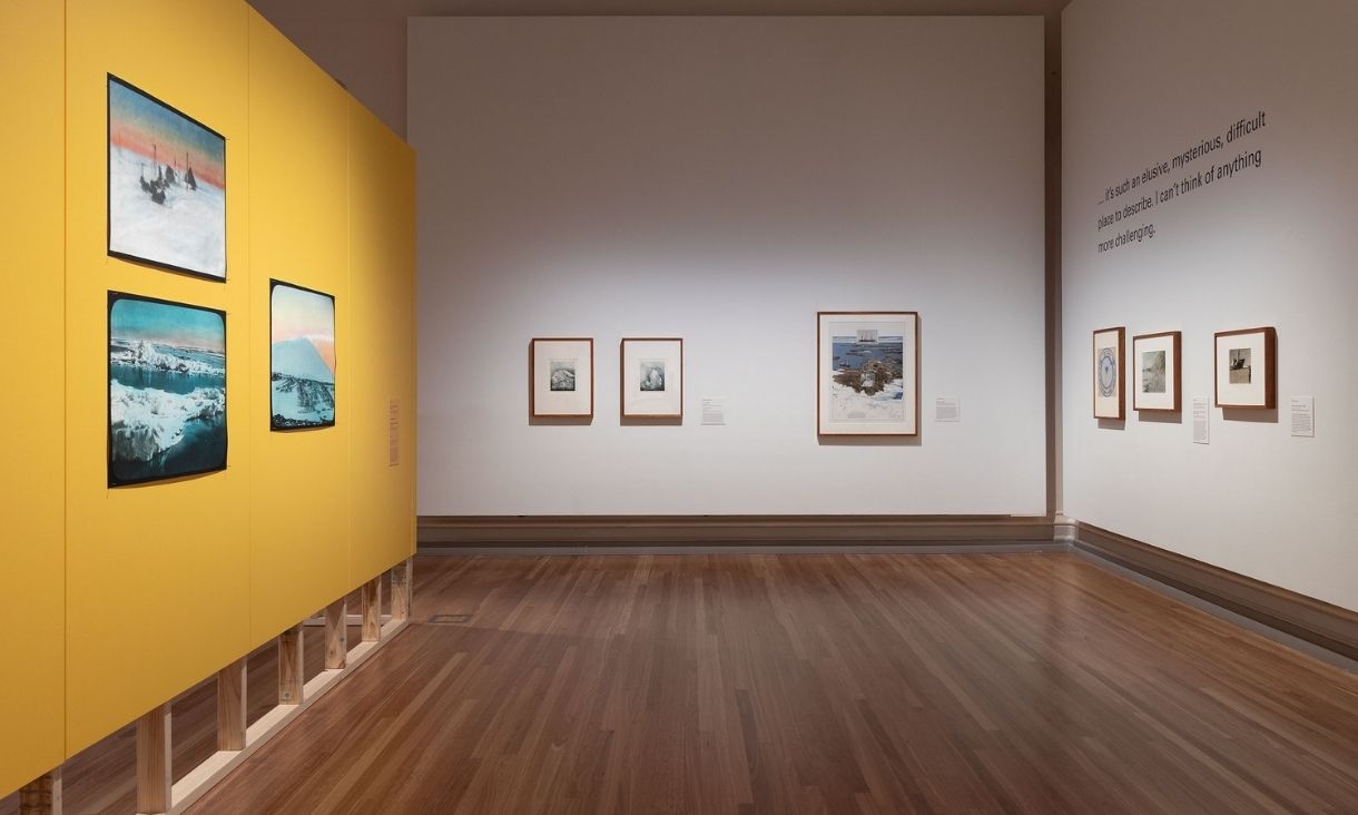 On the left a bright yellow wall, representing the snow machines they use for transport, has three large prints. The back wall and right wall are white with frames lined up across them. 