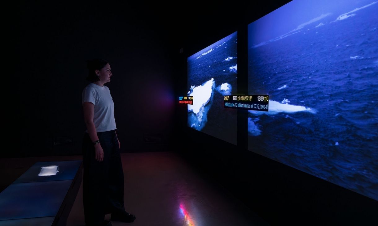 A woman in a white top stands in a very dark room, a bright screen to the right depicts an arctic ocean with icebergs and whitecaps. There is computer-like text on the screen that is undistinguishable.