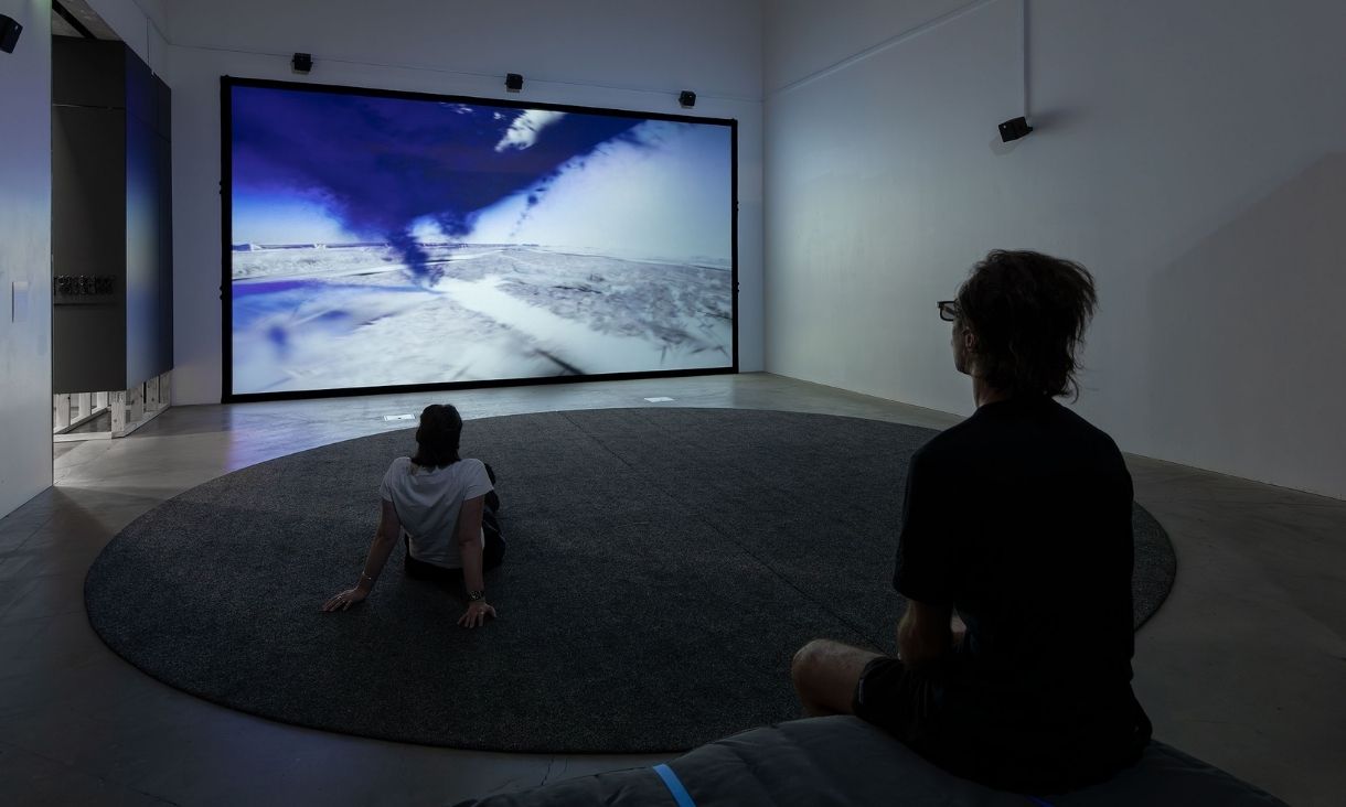 Two people sit in a dark room, one on the floor and the other on a bench, with a huge screen showing the sky and an arctic landscape.