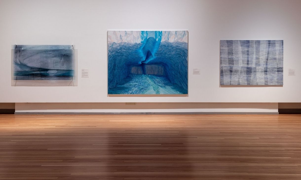 Three very large artworks are lined up on against a white wall, they've got textured blue lines that look soft against warm grey backgrounds.