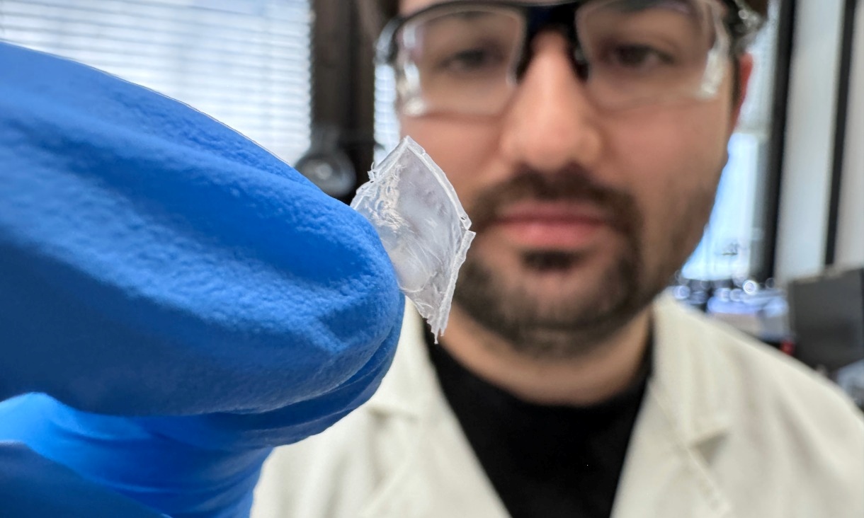 A gloved hand holds a thin piece of nylon film developed as a durable energy‑harvesting device, with laboratory equipment and windows visible in the background.
