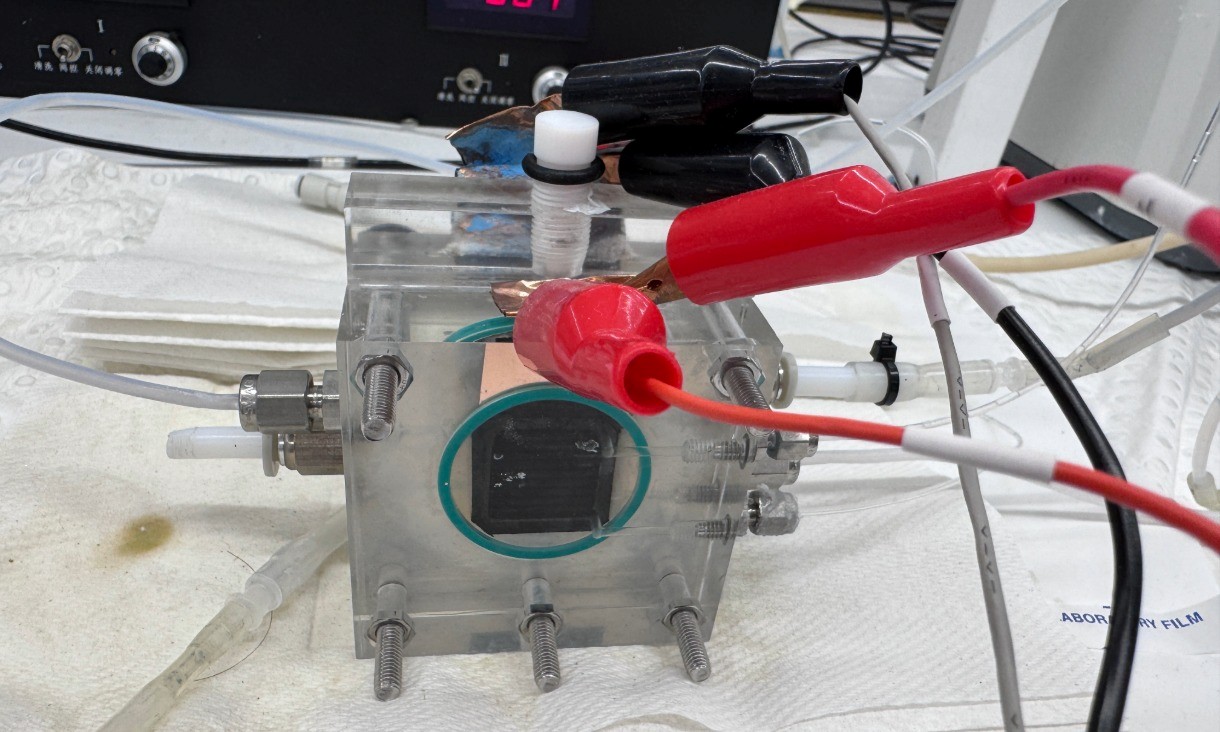 Close up of a small transparent carbon conversion device connected to wires and clamps on a laboratory bench. The device is fitted with bolts and tubing, with additional equipment visible in the background.