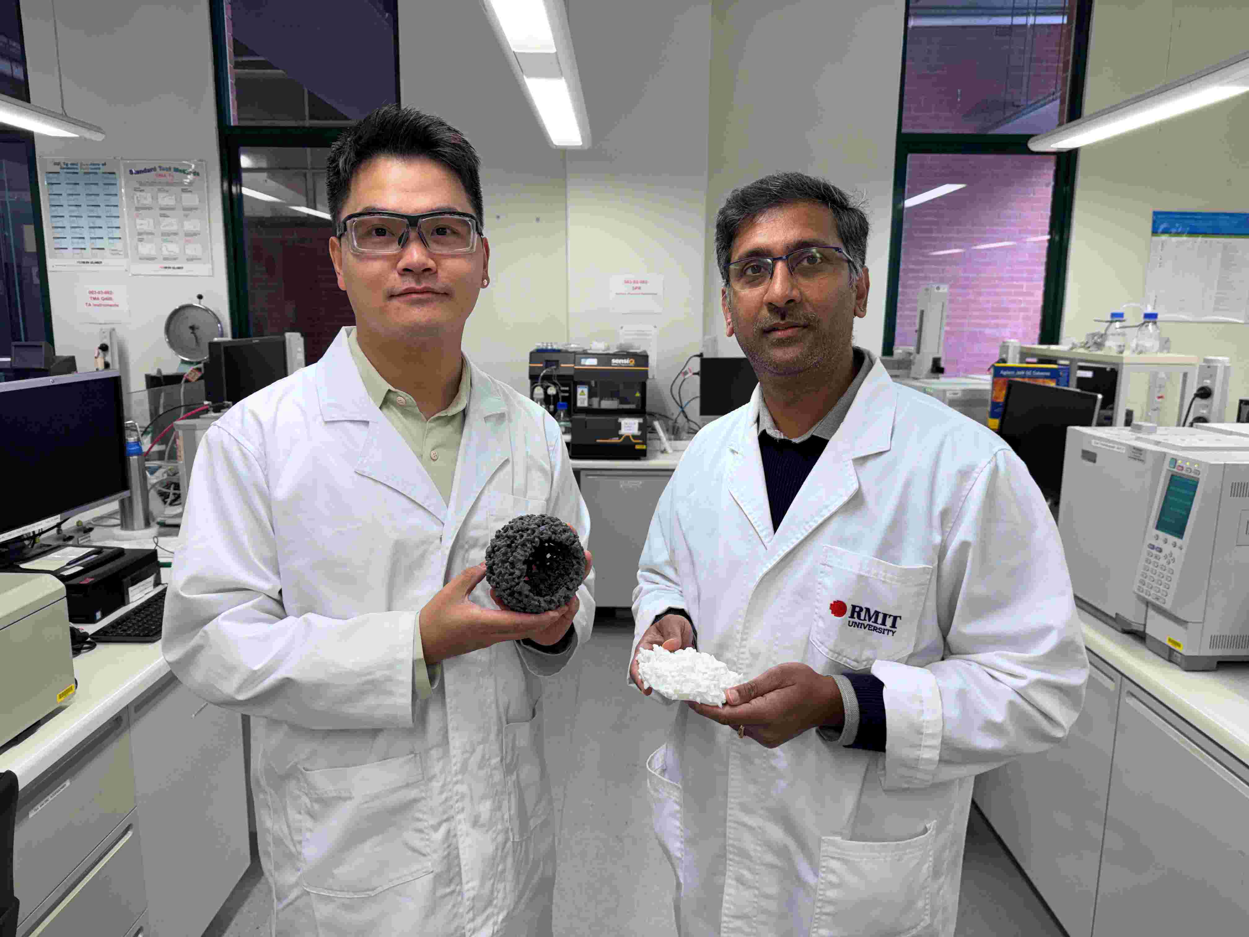 Dr Derek Hao and Associate Professor Ravichandar Babarao stand in an RMIT laboratory surrounded by scientific instruments. Dr Hao holds a grey, porous spherical model of the low-cost material developed in the study.