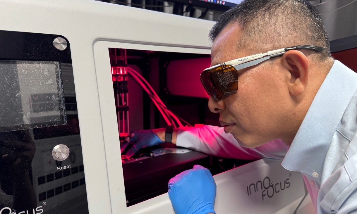 Close up of a researcher wearing protective glasses and gloves working inside a nanofabrication machine illuminated with red light, adjusting components within an enclosed chamber used to build and analyse microscopic structures.