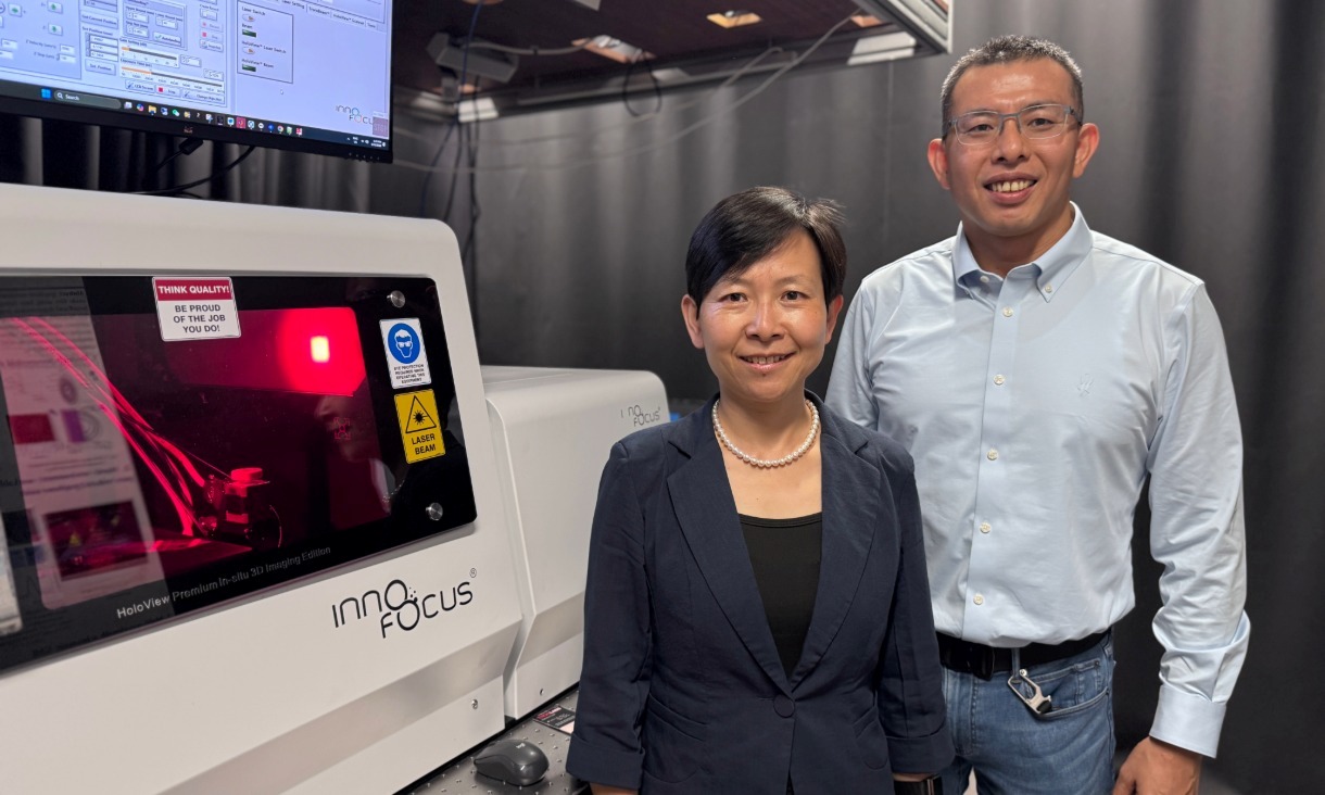 Two researchers standing beside a large laboratory nanofabrication system with a control screen and enclosed chamber in a controlled lab environment.