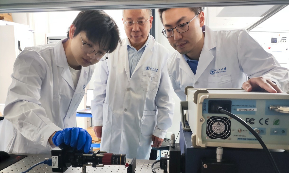 Three researchers in a laboratory setting examining equipment, with lab benches and instruments visible in the background.