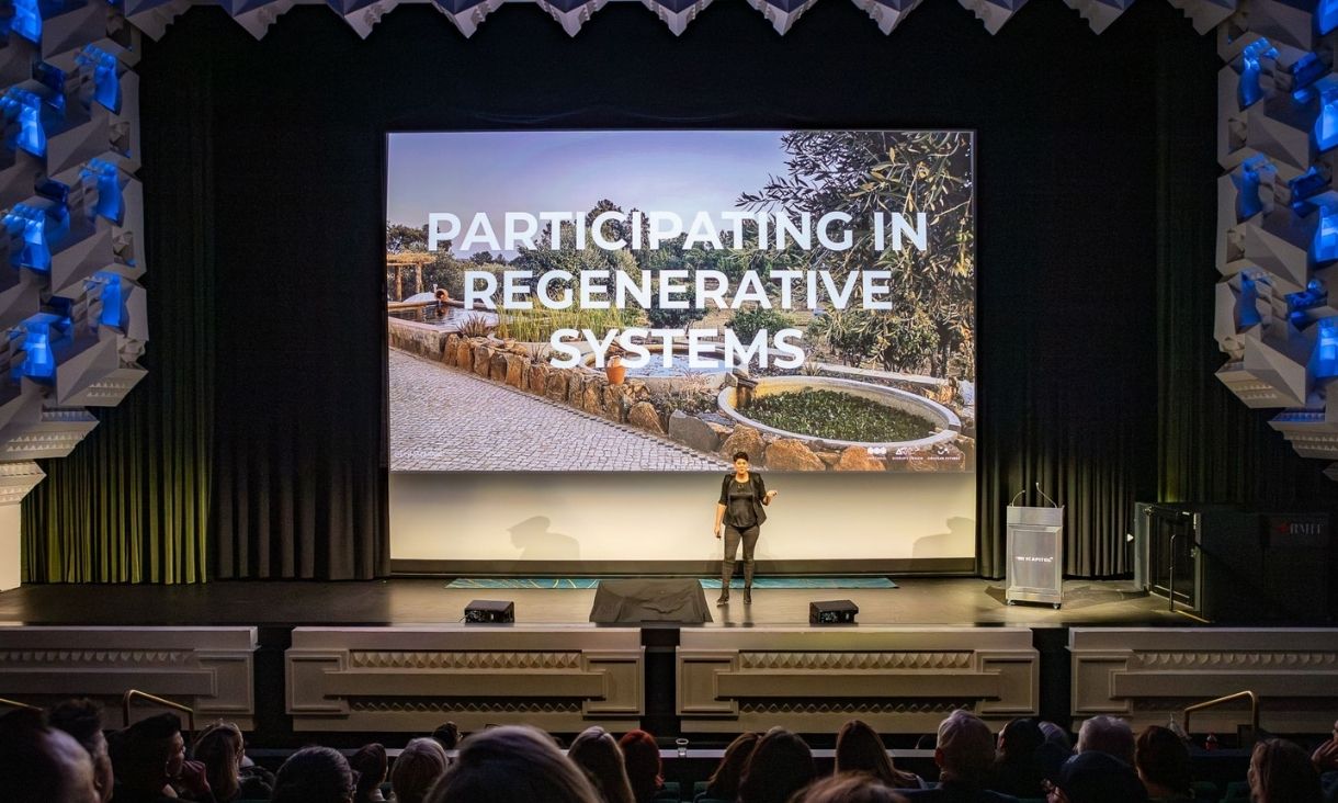 A speaker stands on the Capitol stage, with a giant projection saying 'participating in regenerative systems'.
