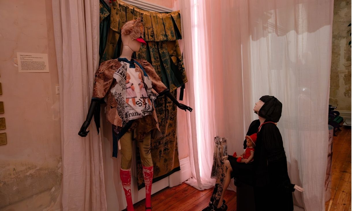 Two mannequins are in a room, one sitting and another standing, there are bright pink lights and soft curtains.