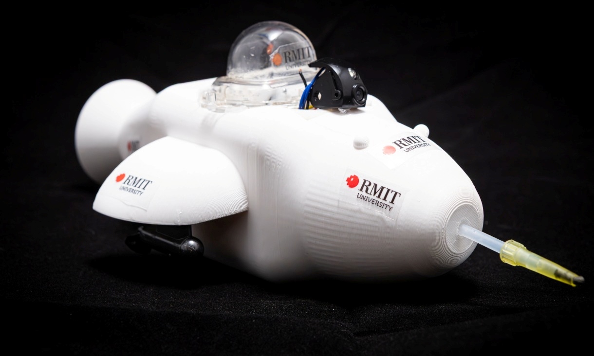 A white, dolphin‑shaped robotic minibot with RMIT University branding sits on a dark surface. The device has a rounded body, a clear dome compartment on top, small side fins and a front nozzle used for drawing in oil during clean‑up tests.