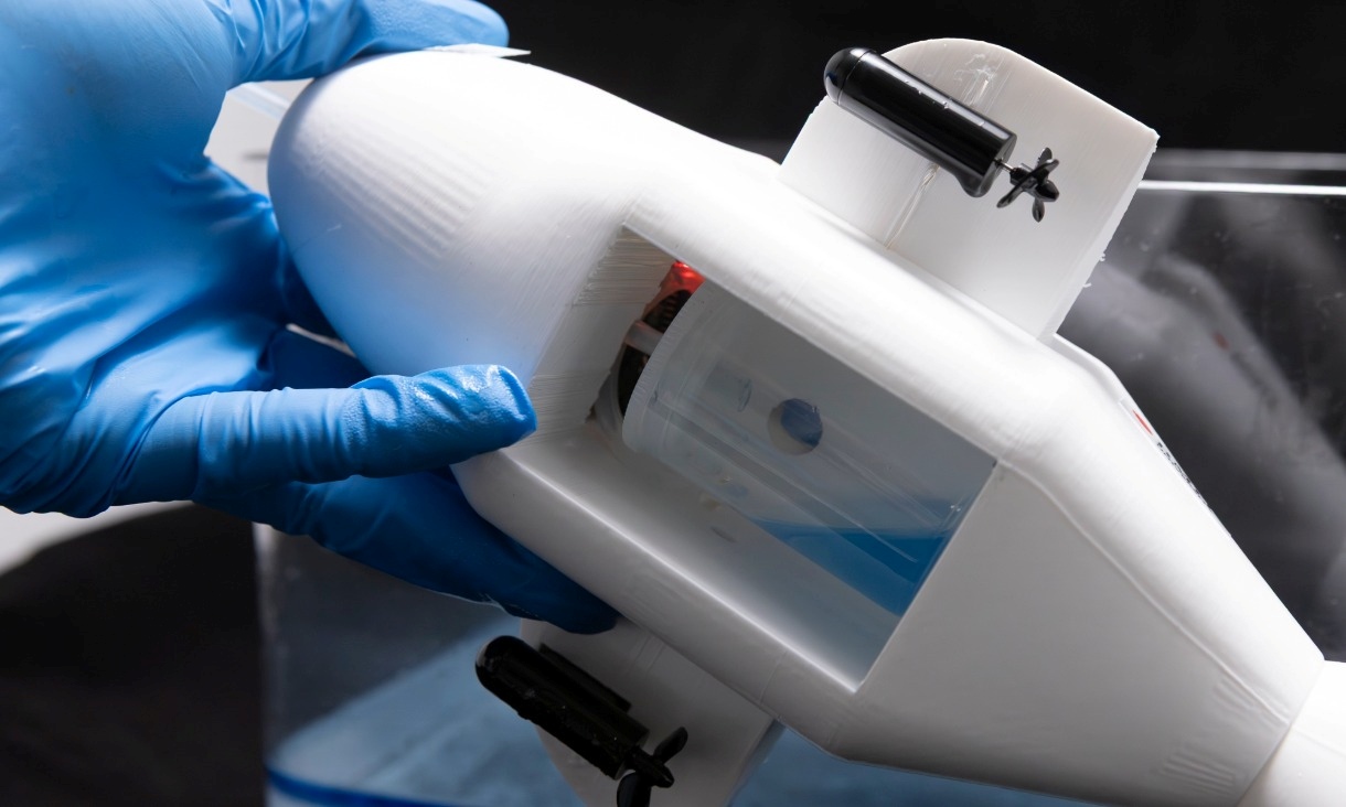 A gloved hand holds a white dolphin‑shaped minibot over a clear tank of water. A cut‑out section on the side of the device shows an internal chamber containing blue kerosene oil collected during testing. The minibot features small black propellers and smooth, streamlined casing.