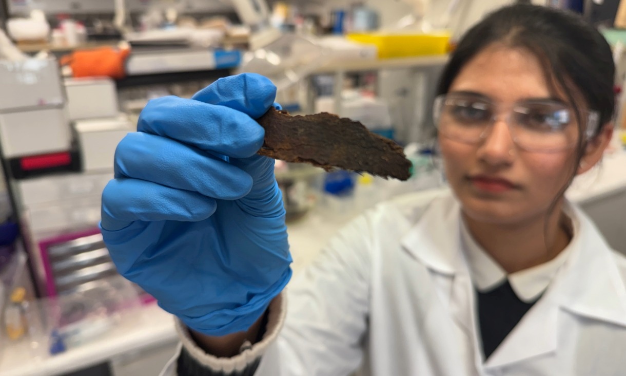 A gloved hand holds up a small, dark, irregularly shaped material sample in the foreground, with a researcher in a laboratory coat visible softly out of focus in the background.