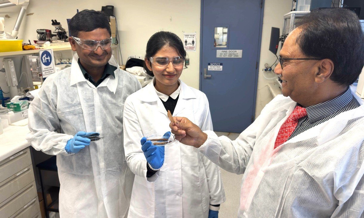 Three researchers wearing laboratory coats and gloves stand in a laboratory and look down at a small, dark sample being held between gloved hands.