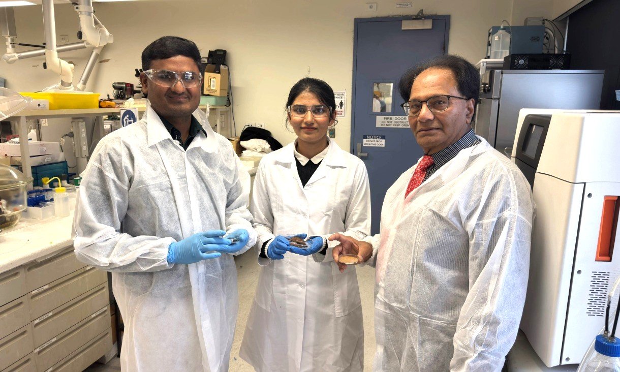 Three researchers wearing laboratory coats stand together in a laboratory. One researcher is positioned on the right, with two colleagues standing to the left and centre, with laboratory benches and equipment visible behind them.