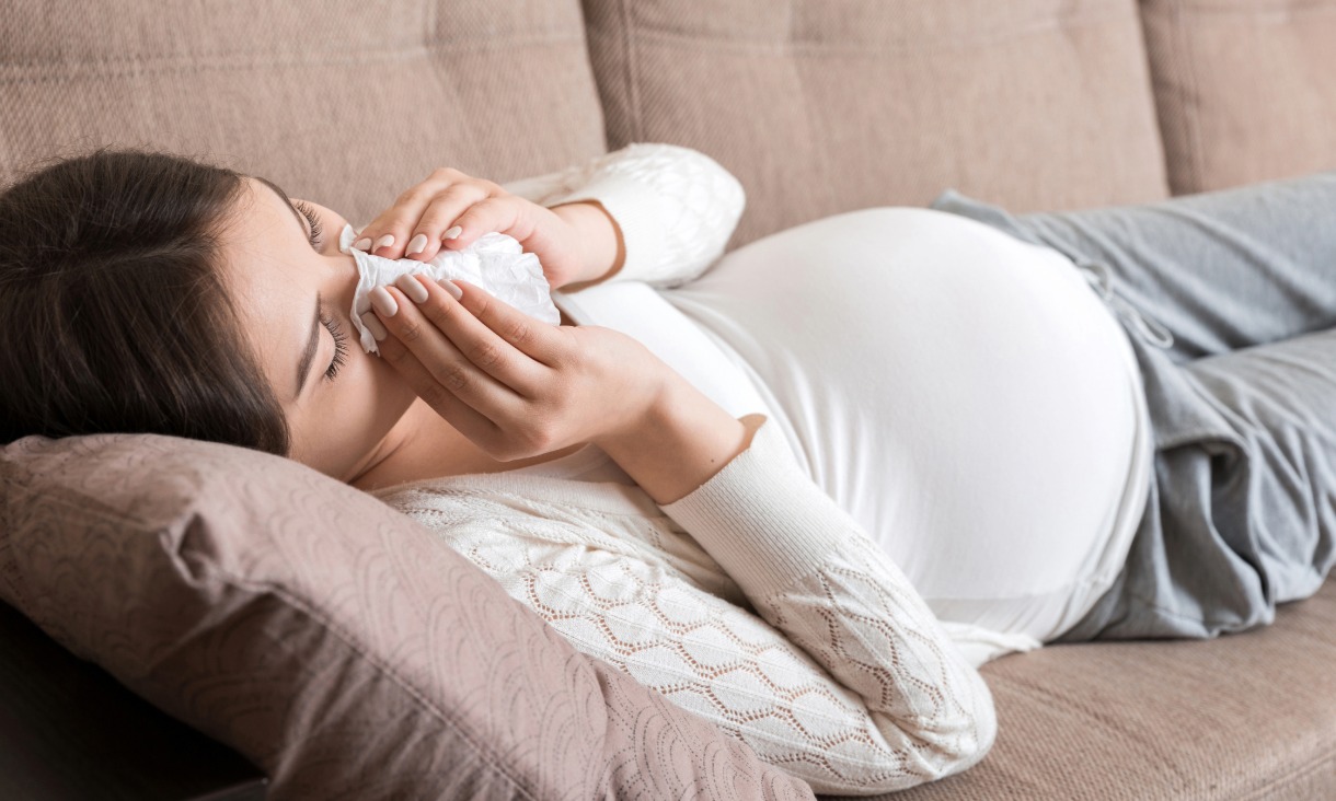 Pregnant woman lying on the couch blowing her nose with a tissue.