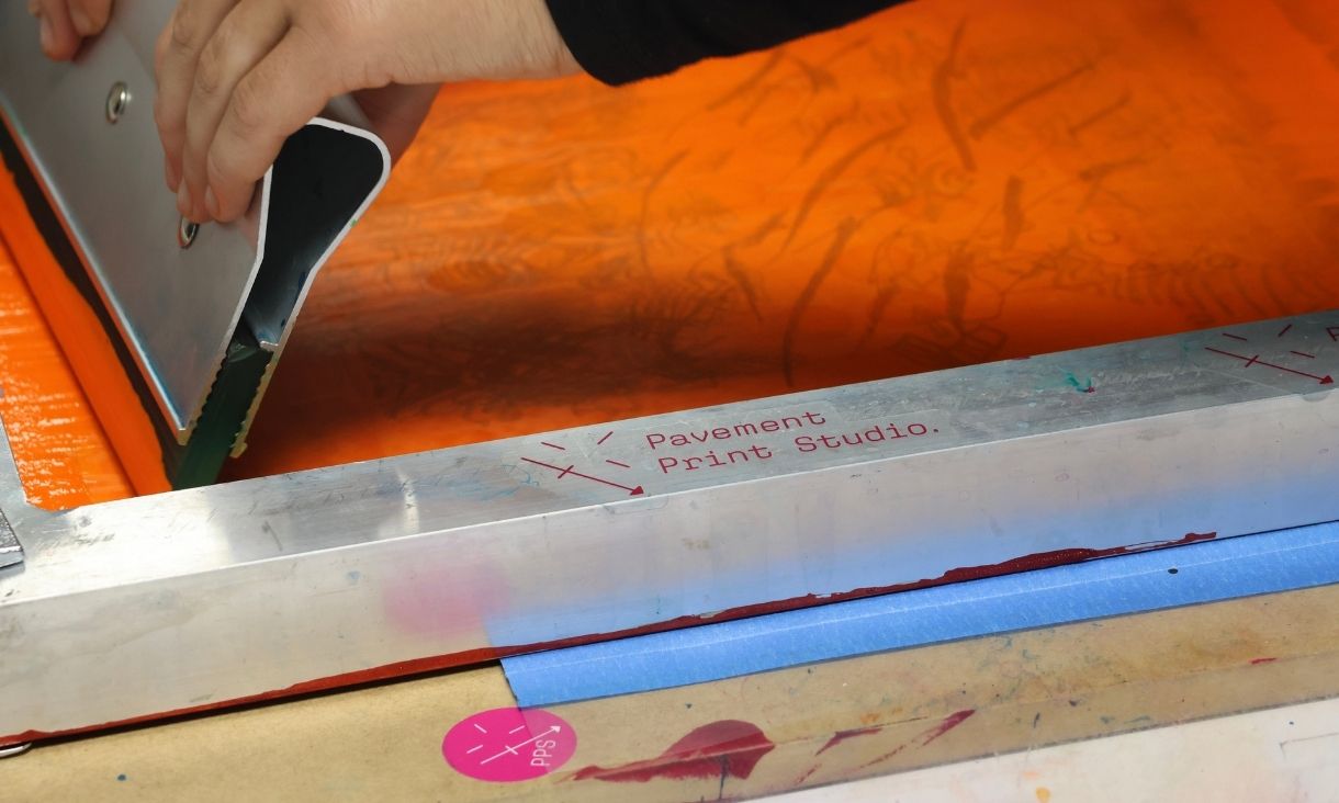 A screen printer with the Pavement Print Studio logo is loaded up with bright orange ink.