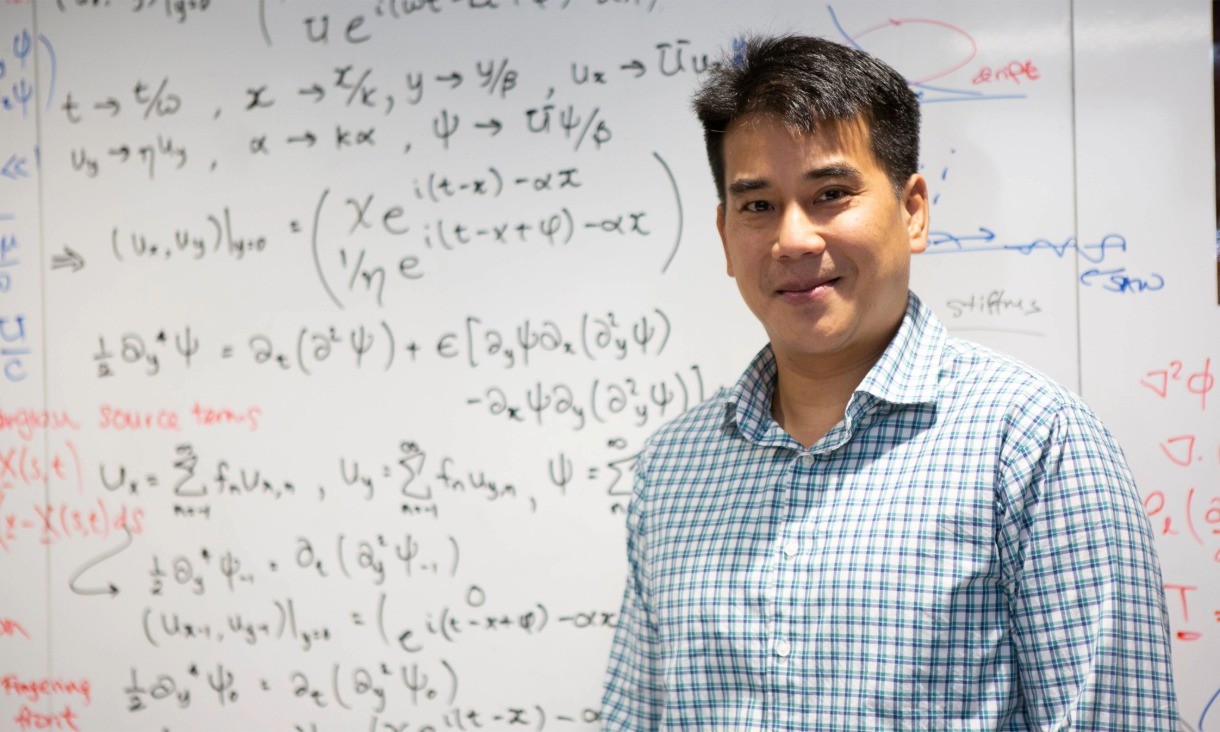 A person wearing a checkered shirt stands in front of a whiteboard covered with handwritten equations and diagrams.
