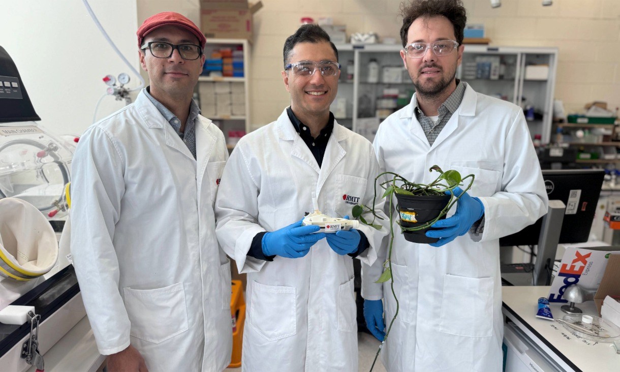Three researchers in lab coats stand side by side in a laboratory surrounded by equipment, posing for a group photograph. The researcher on the right holds a potted plant while the researcher in the middle holds a sound wave nebuliser with the RMIT logo.