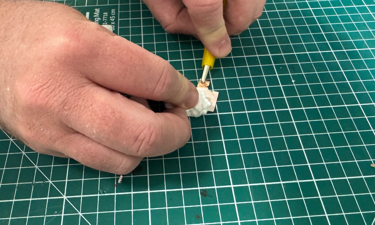 Two hands on a green cutting mat. One presses a small polystyrene block onto a piece of copper foil, while the other touches a yellow-handled electrode to the foil.