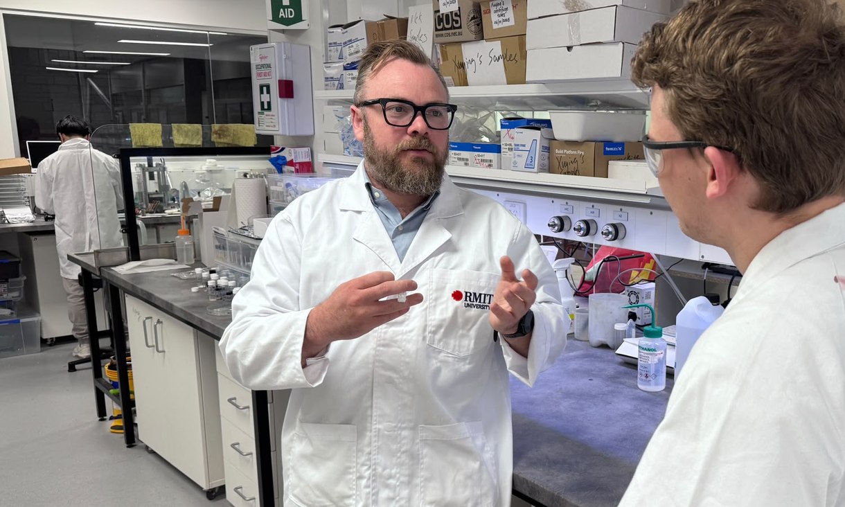 Dr Peter Sherrell, wearing glasses and an RMIT lab coat, gestures as he talks with a colleague in a laboratory.