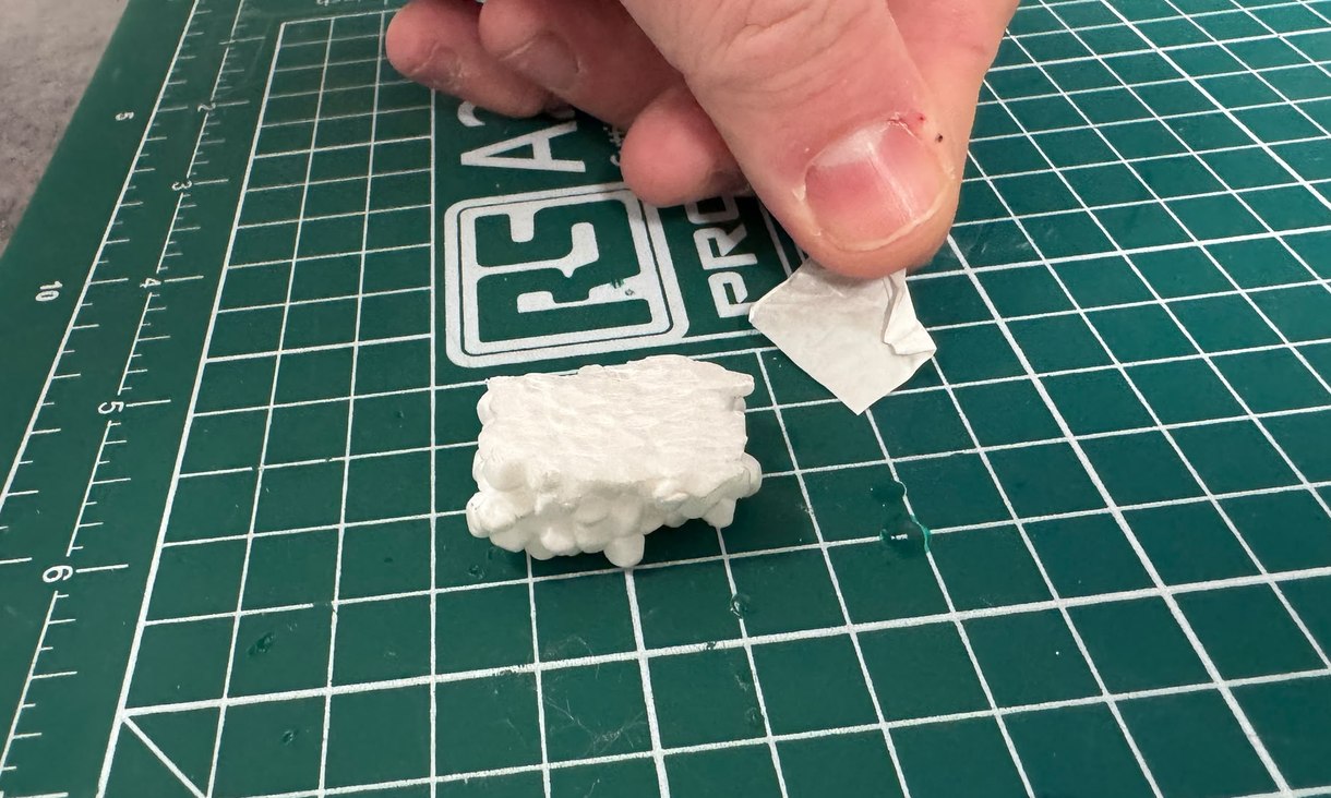 A hand holds a thin white polystyrene layer just above a small polystyrene block on a green cutting mat.