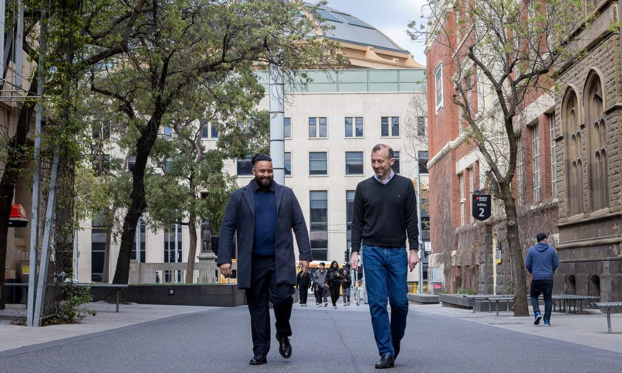 Two men walking along a road with buildings and trees and other people in the background.